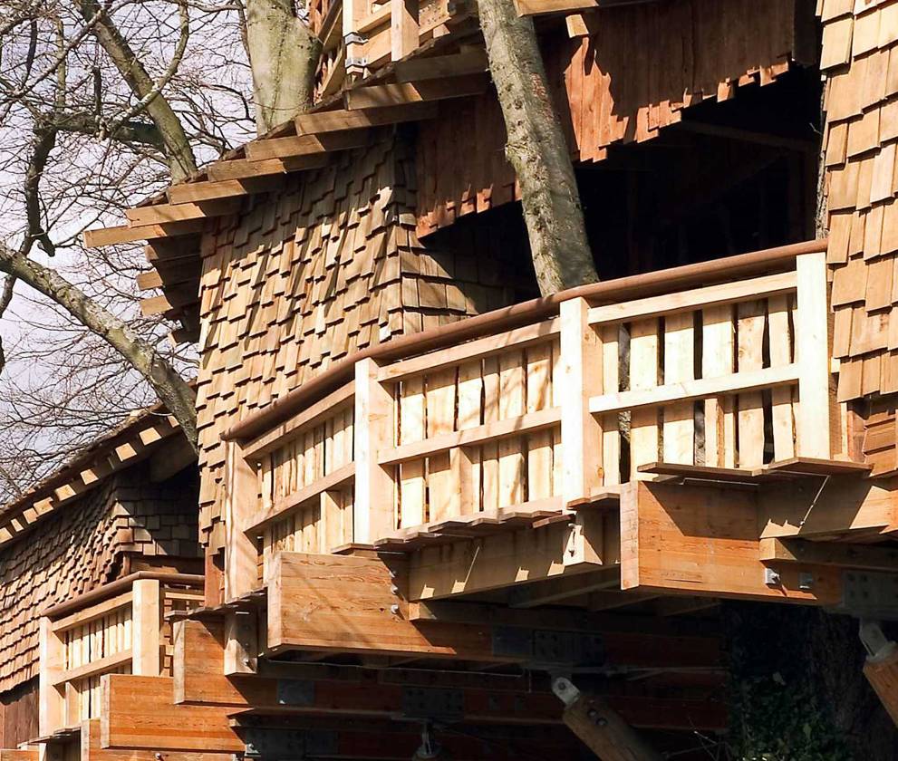 A large wooden treehouse, supported by timber beams, nestled among semi-mature trees in a fairytale-like setting.