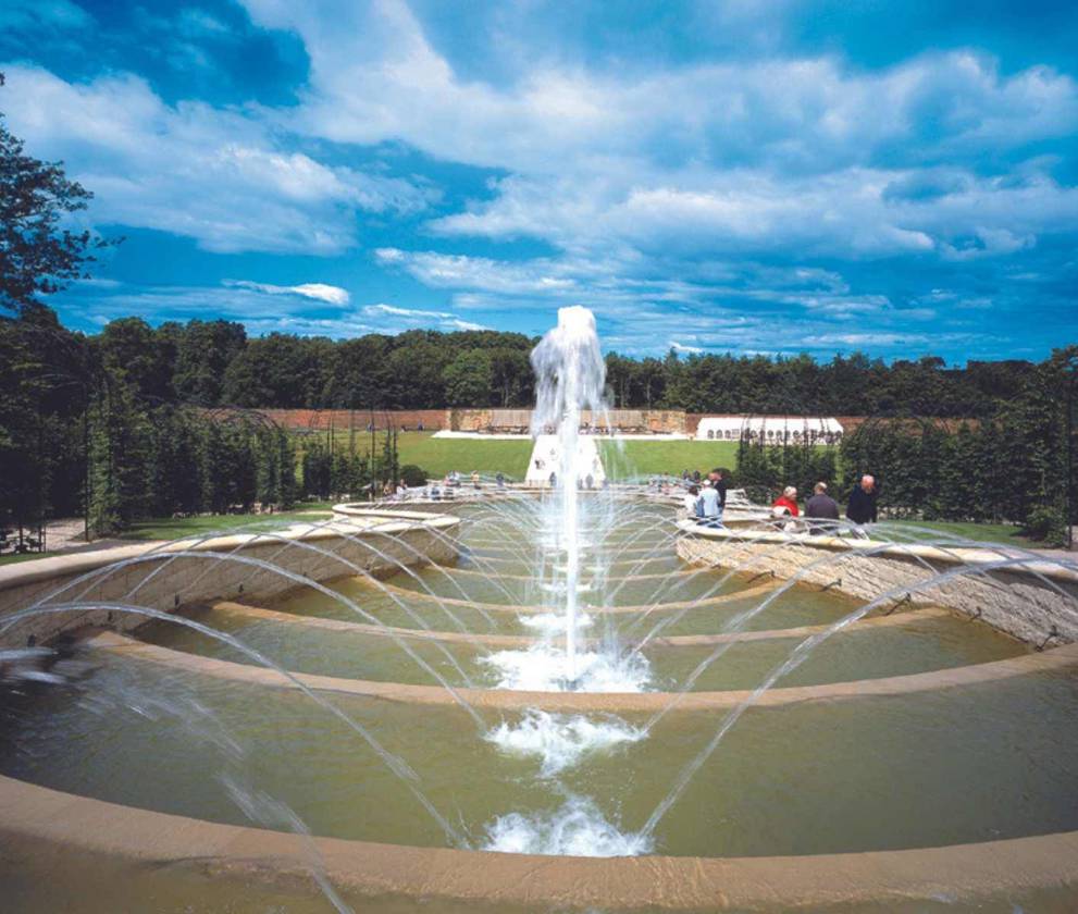 A wide shot of the Grand Cascade fountain with water flowing down multiple tiers, surrounded by lush greenery.